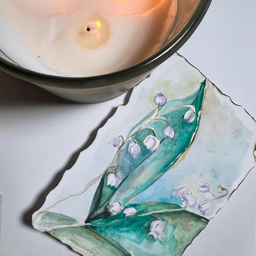 Watercolor of lily of the valley with gold accents on torn paper beside a lit candle